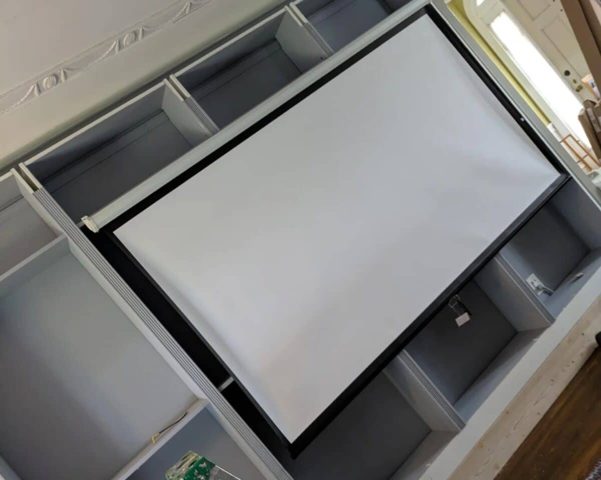 Projection screen mounted in a room with gray shelves, viewed at an angle from the side window.