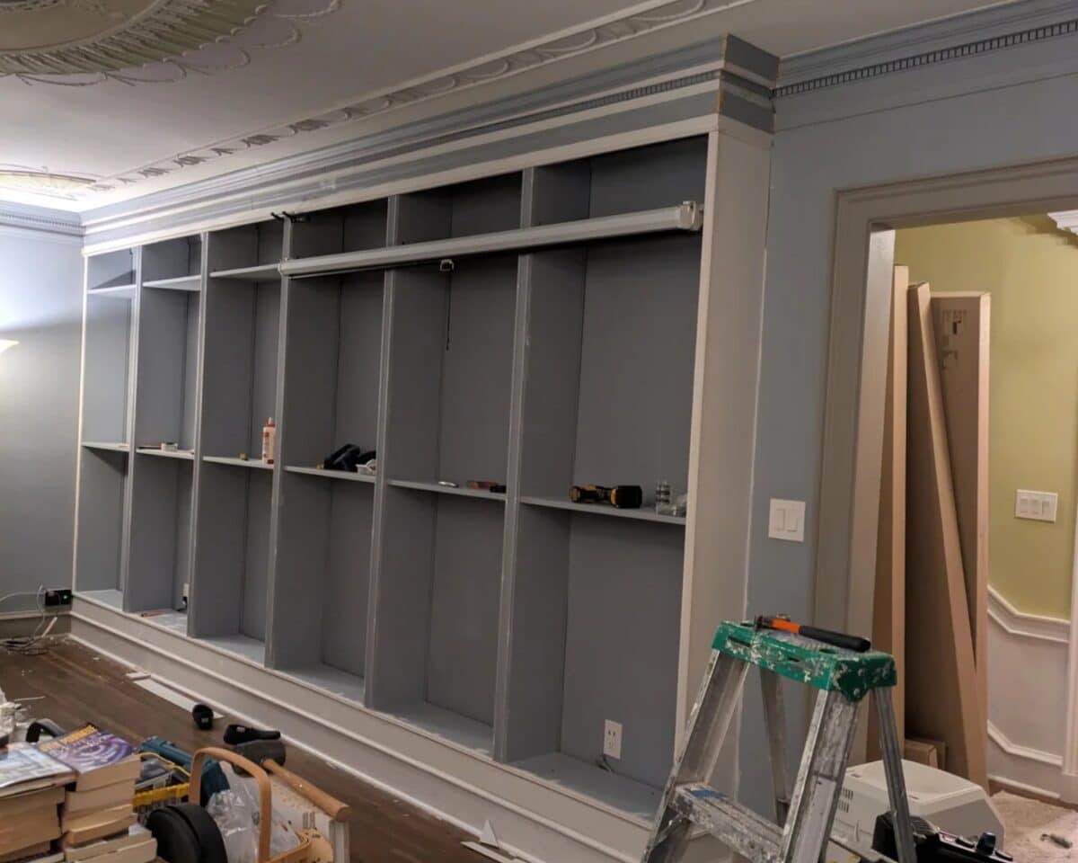 Grey built-in shelves along a wall being installed, with a step ladder and tools in a room under renovation, ceiling molding visible.