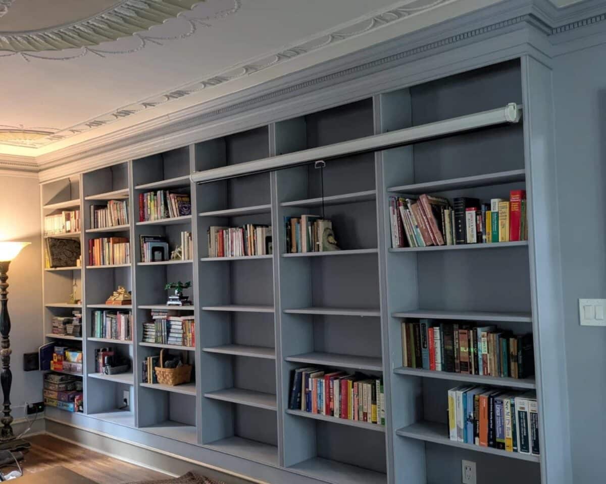 Blue-gray IKEA BILLY built-in bookshelves filled with books and a few decorative items along a wall in a living room with decorative crown molding. Projector screen installed along one level of shelves.
