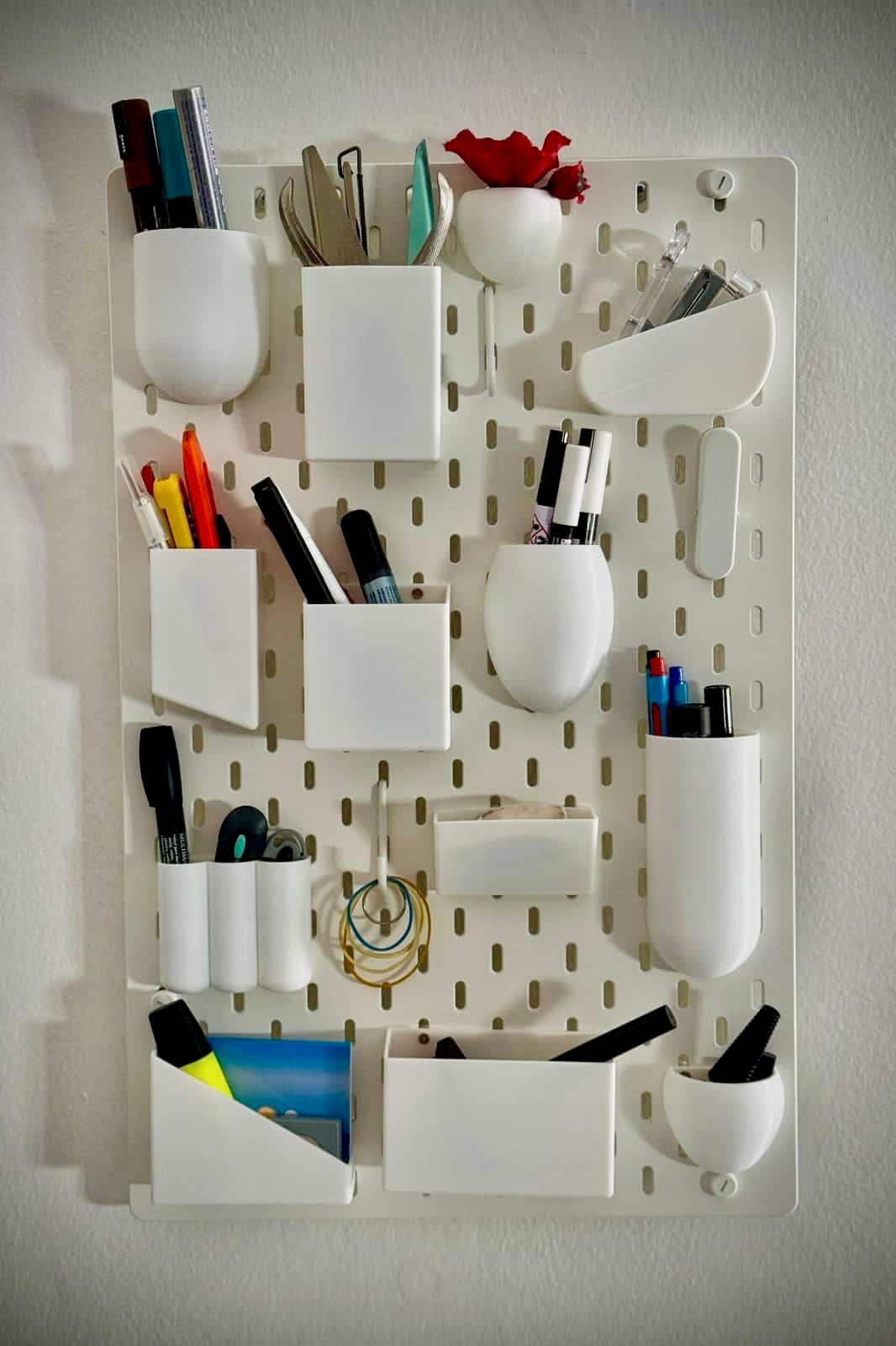 Uten IKEA Skadis - IKEA Hackers IKEA SKÅDIS pegboard UTEN wall organizer inspired hack. White pegboard organizer mounted on wall with multiple white containers holding markers, scissors, pens, and small office supplies, plus a red artificial flower in a white vase at the top.