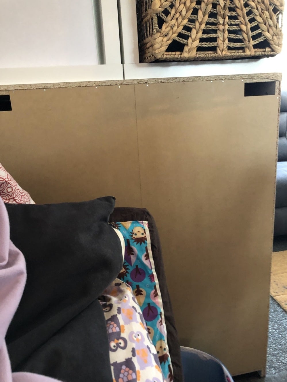 IKEA MALM dresser backing board which is a large brown panel or board propped upright behind a couch with colorful pillows in the foreground, indoors.