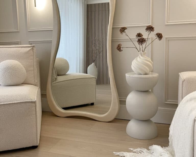 blanda-matt-table-in-room-768x614 - IKEA Hackers Neutral contemporary living room featuring a tall curved-framed mirror, light wood floors, and a stacked pedestal with a white vase and dried flowers beside a tufted seating area.