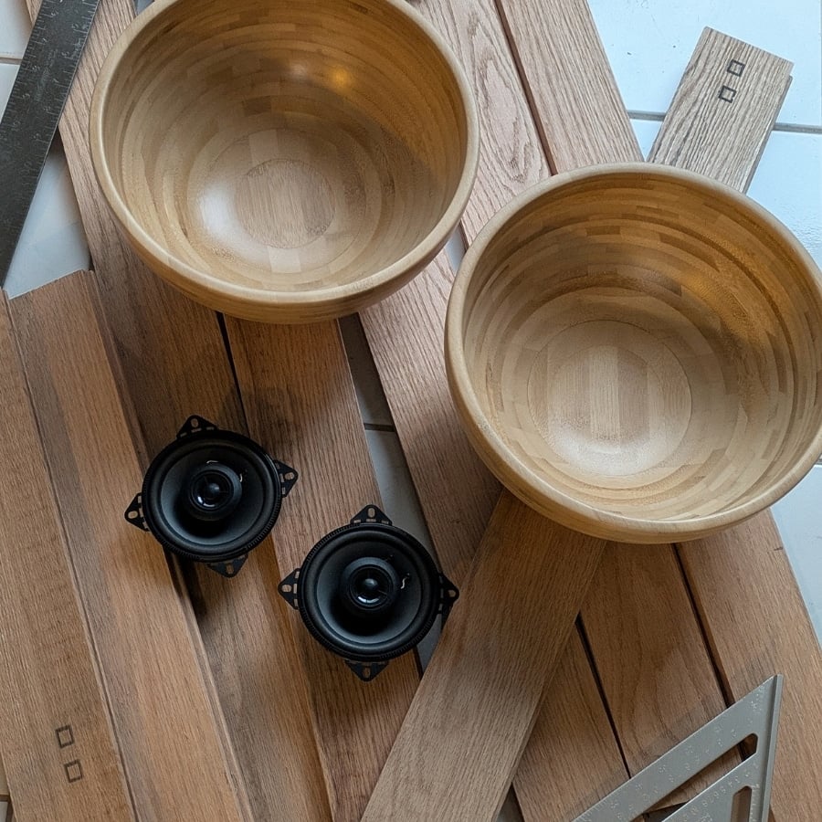 44 - IKEA Hackers Close-up of a woodworking workspace showing two light wooden bowls, wooden planks, a ruler, a metal square, and two small round black speaker drivers.