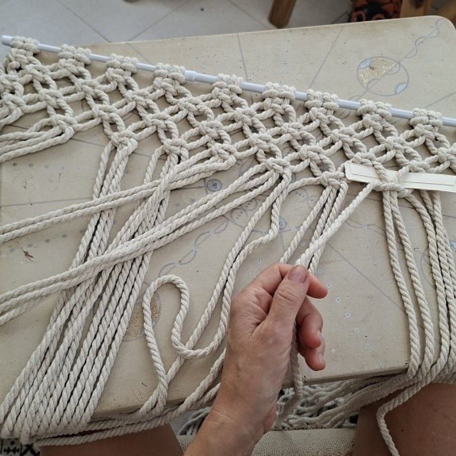 Close-up of hands knotting off-white rope into a macrame pattern on a worktable with a rod running through the top edge.