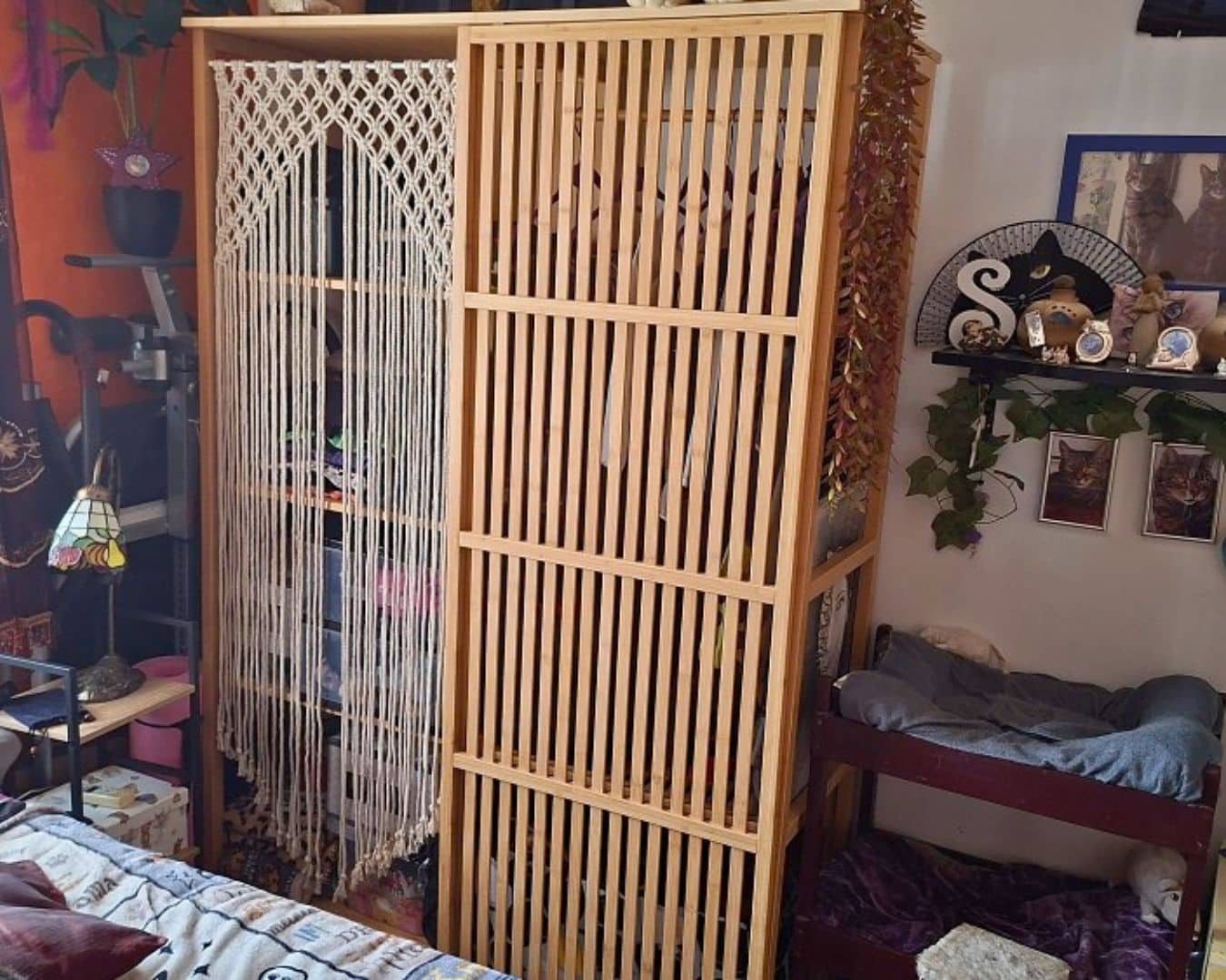 Wooden slatted wardrobe with a macrame curtain on the left in a cluttered bedroom corner.